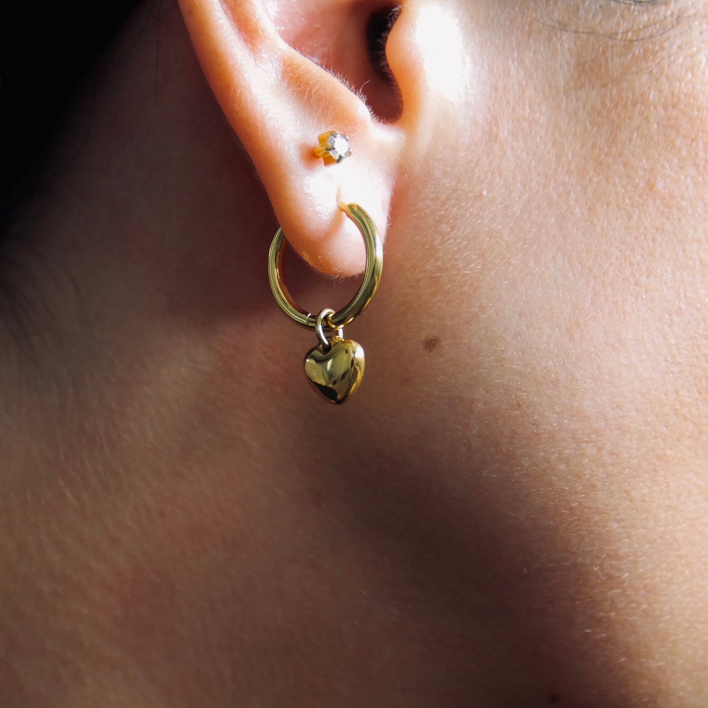 Close-up of a gold hoop earring with a heart charm worn by a person.
