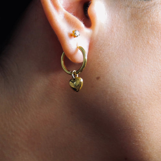 Close-up of a gold hoop earring with a heart charm worn by a person.